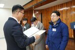 2월 정례조회에 정장을 입고 안경을 낀 남성이 밝은 파란색 두꺼운 패딩을 입은 김남우에게 패를 수여하고 있다.