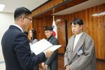 2월 정례조회에 정장을 입고 안경을 낀 남성이 흰색 목티에 밝은 오트밀색 코트를 입은 최성관에게 패를 수여하고 있다.